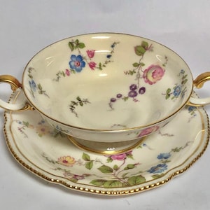 May include: A vintage porcelain soup bowl and saucer set with a floral pattern in shades of pink, blue, and yellow. The set has a gold trim and a scalloped edge.