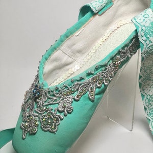 May include: A pair of turquoise ballet shoes decorated with silver lace, sequins, and rhinestones. The shoes are on a clear acrylic stand.