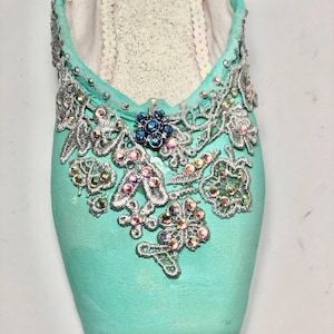 May include: A pair of turquoise ballet shoes decorated with silver lace and iridescent rhinestones. The shoes have a pointed toe and a delicate design.