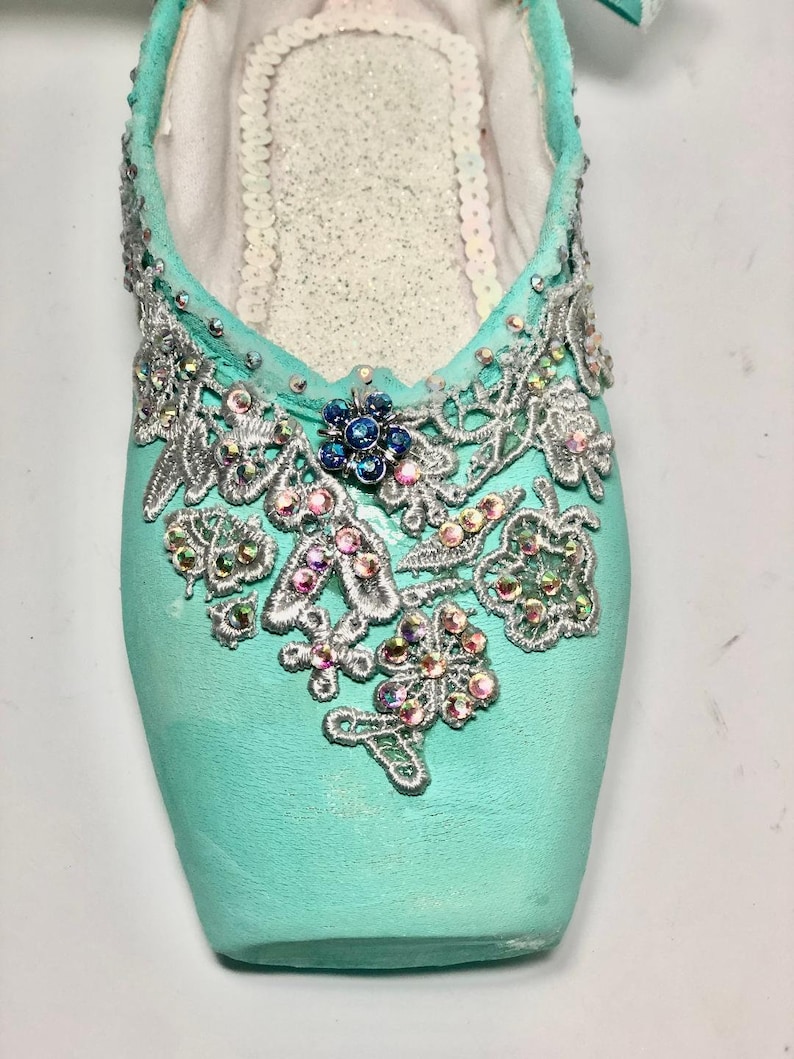 May include: A pair of turquoise ballet shoes decorated with silver lace and iridescent rhinestones. The shoes have a pointed toe and a delicate design.