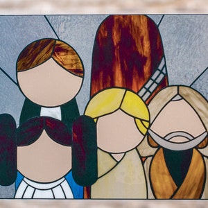 Puede incluir: Una ventana de vitrales que representa a tres figuras. Las figuras son una mujer con cabello castaño, una niña con cabello rubio y un hombre con barba. La mujer lleva un vestido azul, la niña lleva un vestido blanco y el hombre lleva una túnica marrón. El fondo es azul claro.