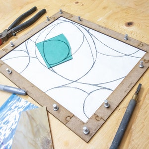 May include: A stained glass project in progress, with a wooden frame holding a piece of glass with a design drawn on it. A piece of green glass is placed on top of the design. A silver marker, a glass cutter, and a pair of pliers are also visible.