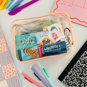 以下が含まれることがあります： Tide、Liquid I.V.、Dr. Nmkのパッケージなど、様々なアイテムが入った、透明で長方形のピーチ色の縁取りのポーチ。ポーチの周りには、カラフルなペン、ノート、花柄のメモ帳が置かれています。