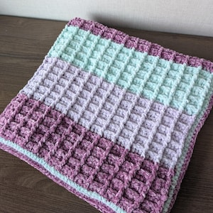 Puede incluir: Una suave manta de bebé tejida a crochet con un patrón de punto de waffle en tonos lavanda, verde claro y rosa.