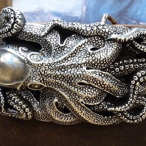 May include: A silver-colored octopus belt buckle with intricate detailing. The buckle features a three-dimensional design of an octopus with its tentacles wrapped around the buckle. The buckle is attached to a brown leather belt.