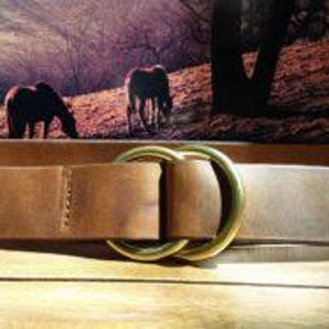 Puede incluir: Un cintur&oacute;n de cuero marr&oacute;n con una hebilla dorada de doble lazo. El cintur&oacute;n se muestra sobre una superficie de madera, con una imagen enmarcada de caballos en el fondo. El cintur&oacute;n mide aproximadamente 3,8 cm de ancho.