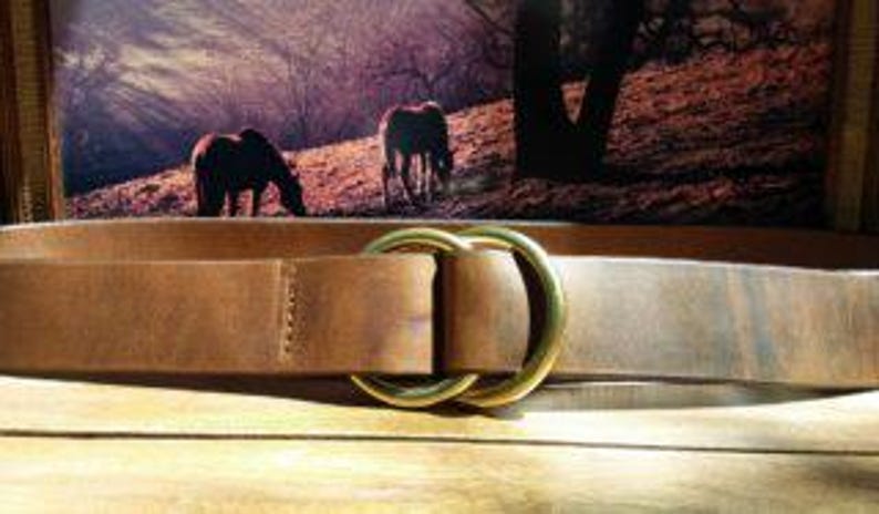 Puede incluir: Un cintur&oacute;n de cuero marr&oacute;n con una hebilla dorada de doble lazo. El cintur&oacute;n se muestra sobre una superficie de madera, con una imagen enmarcada de caballos en el fondo. El cintur&oacute;n mide aproximadamente 3,8 cm de ancho.