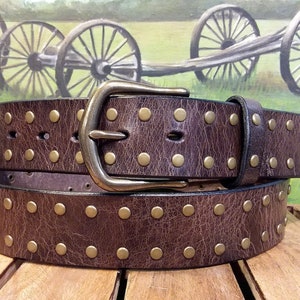May include: A dark brown leather belt featuring a distressed, textured finish and two parallel rows of small, round, brass-toned metal studs. The belt is secured with a matching brass-toned metal prong buckle and includes a single leather keeper loop.