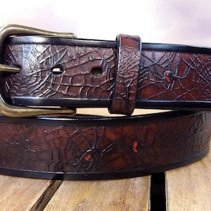 Op de afbeelding: Een donkerbruine leren riem met een reliëfpatroon van spinnenwebben over de hele band. De riem bevat kleine rode accenten op de spinnen en is afgewerkt met een messingkleurige metalen gesp en een bijpassende leren lus.