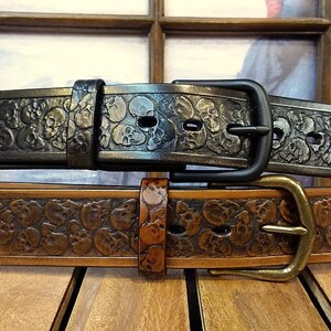 以下が含まれることがあります： ストラップ全体にスカルのエンボス加工が施された2本のレザーベルト。上のベルトはブラックでマットブラックのバックルが付いており「Black Antique」と表記されています。下のベルトはタンカラーでアンティーク真鍮のバックルが付いており「Tan Antique」と表記されています。