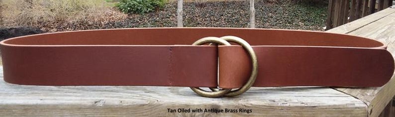 Puede incluir: Cintur&oacute;n de cuero curtido de color canela con anillos de lat&oacute;n antiguo. El cintur&oacute;n tiene una hebilla de doble anillo. El texto "Tan Oiled with Antique Brass Rings" es visible en la parte inferior de la imagen. Dise&ntilde;o cl&aacute;sico.