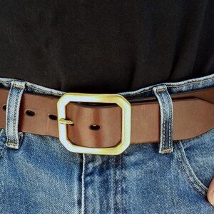 Op de afbeelding: Een bruine leren riem met een goudkleurige vierkante gesp wordt over een spijkerbroek gedragen. De riem heeft een klassiek ontwerp met een eenvoudige, elegante uitstraling. De riem is ongeveer 3,8 cm breed.