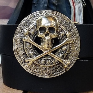 May include: A gold-toned belt buckle with an ornate design featuring a skull and crossbones. The skull has a detailed face with eye sockets and a prominent nose.