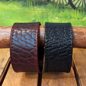 May include: Two wide leather cuff bracelets with a textured, pebbled surface. One bracelet is deep burgundy and the other is black. Both cuffs feature visible stitching along the edges and are displayed side by side on a wooden branch.