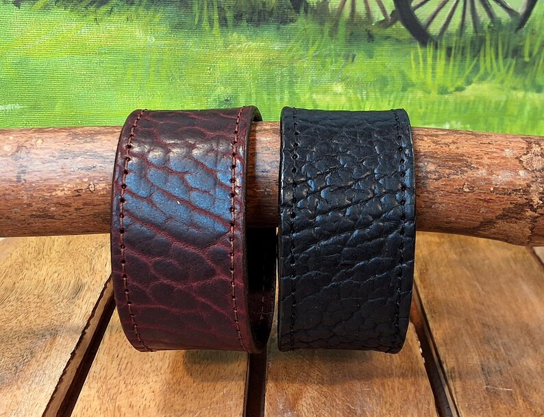 May include: Two wide leather cuff bracelets with a textured, pebbled surface. One bracelet is deep burgundy and the other is black. Both cuffs feature visible stitching along the edges and are displayed side by side on a wooden branch.
