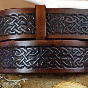 May include: A brown leather belt with a Celtic knot design. The belt is embossed with a dark brown pattern.
