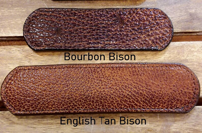 May include: Two rectangular leather strips with rounded ends and a textured surface. The top strip is dark brown with the text Bourbon Bison below it. The bottom strip is a lighter tan color with the text English Tan Bison below it.