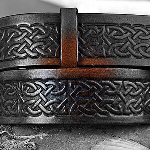 May include: A black leather belt with an intricate Celtic knot design embossed on the surface. The belt has a brown leather buckle.