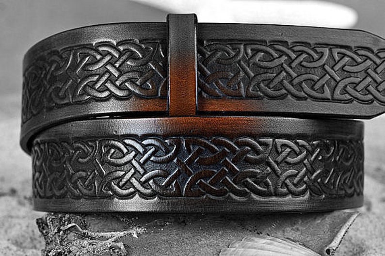 May include: A black leather belt with an intricate Celtic knot design embossed on the surface. The belt has a brown leather buckle.