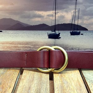 Puede incluir: Un cintur&oacute;n de cuero burdeos con una hebilla de doble anillo dorado. El cintur&oacute;n se muestra sobre un fondo de paisaje marino con veleros y un cielo nublado. El cintur&oacute;n est&aacute; sobre una superficie de madera.