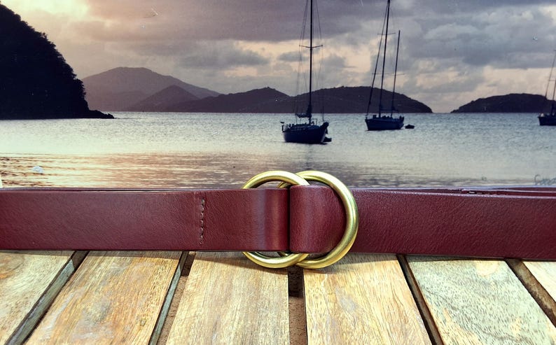 Puede incluir: Un cintur&oacute;n de cuero burdeos con una hebilla de doble anillo dorado. El cintur&oacute;n se muestra sobre un fondo de paisaje marino con veleros y un cielo nublado. El cintur&oacute;n est&aacute; sobre una superficie de madera.
