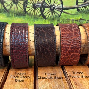 Op de afbeelding: Vijf brede leren armbanden met een getextureerd oppervlak, weergegeven in de kleuren Tucson Black Bison, Black Cherry Bison, Chocolate Bison, Peanut Bison en Cognac Bison, elk voorzien van zichtbare stiksels langs de randen.