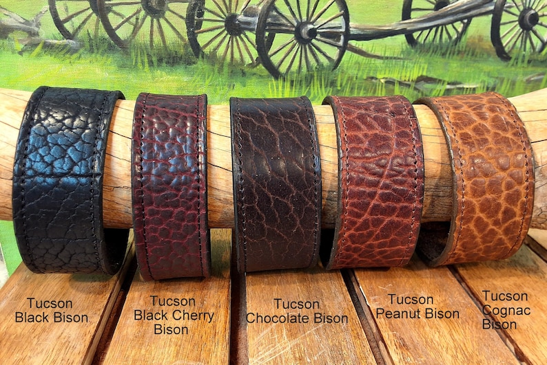 May include: Five wide leather cuff bracelets with a textured surface, displayed in colors labeled as Tucson Black Bison, Black Cherry Bison, Chocolate Bison, Peanut Bison, and Cognac Bison, each featuring visible stitching along the edges.