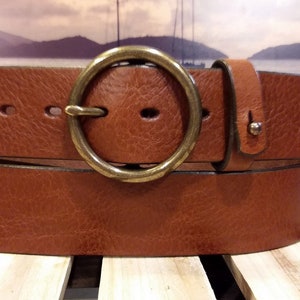 May include: A brown leather belt with a textured surface and a large, circular, antique brass-toned metal buckle. The belt features a single leather keeper loop and multiple oval-shaped adjustment holes along the strap for a secure fit.