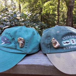 May include: Two teal baseball caps with embroidered designs. One features a whale graphic and the words "Coastal Studies." The other displays "St. Thomas U.S. Virgin Island" with shark and "S" graphics. Both have a decorative oyster.