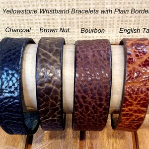 May include: Four Yellowstone wristband bracelets with a plain border, displayed in charcoal, brown nut, bourbon, and English tan colors. Each bracelet features a distinct textured surface and is arranged horizontally on a cylindrical support.