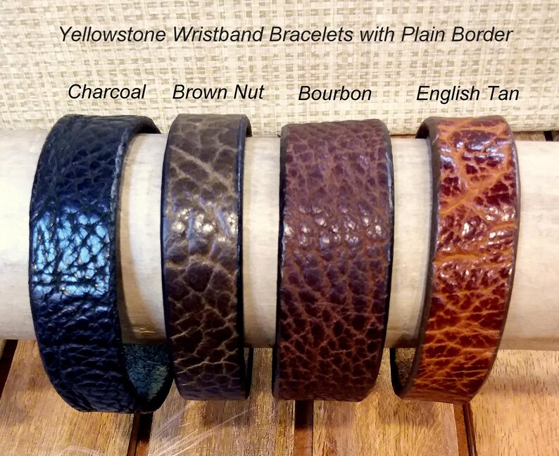 May include: Four Yellowstone wristband bracelets with a plain border, displayed in charcoal, brown nut, bourbon, and English tan colors. Each bracelet features a distinct textured surface and is arranged horizontally on a cylindrical support.