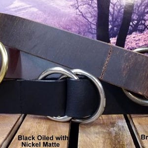 Puede incluir: Tres cinturones de cuero en marr&oacute;n y negro, cada uno con una hebilla de doble anilla. Los cinturones est&aacute;n etiquetados: "Brown Oiled with Natural Brass", "Black Oiled with Nickel Matte" y "Brown Distressed with Antique Brass".