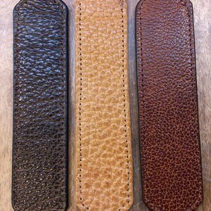 May include: Three rectangular bison leather strips with rounded ends and stitched edges, labeled Yellowstone Bison Leather Colors. The colors are Nut Brown, Natural, and English Tan, each featuring a distinct pebbled texture and grain.