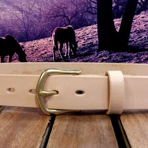 May include: A light brown leather belt with a brass buckle. The belt is on a wooden surface.