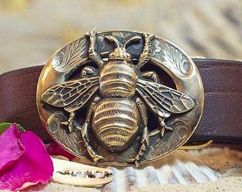 May include: An oval-shaped brass belt buckle featuring a detailed relief of a bee in the center, set against a dark, textured background with engraved leaf patterns along the rim, attached to a smooth, dark brown leather belt strap.
