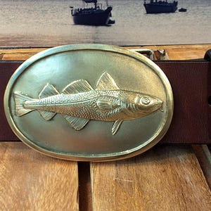 May include: A brown leather belt with a brass buckle featuring a detailed embossed image of a fish. The buckle is oval shaped.