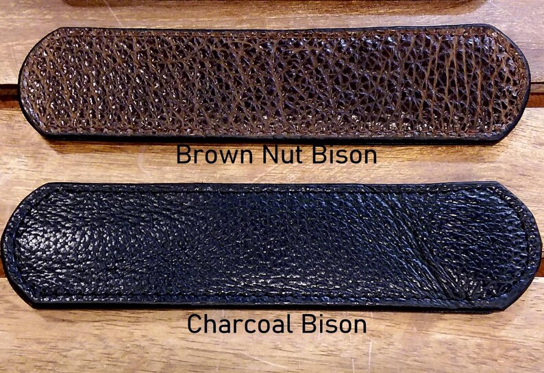 May include: Two rectangular leather strips with rounded ends and stitched edges. The top strip is textured brown leather labeled Brown Nut Bison. The bottom strip is textured black leather labeled Charcoal Bison. Both items feature a pebbled surface.