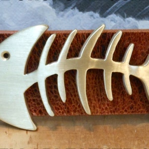 May include: A silver-colored fish bone belt buckle with a detailed design. The buckle is attached to a brown leather belt.