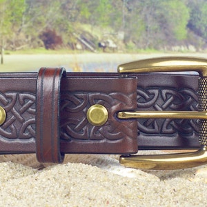 May include: A brown leather belt with a brass buckle and Celtic knot design. The belt has a textured surface and is decorated with brass rivets.