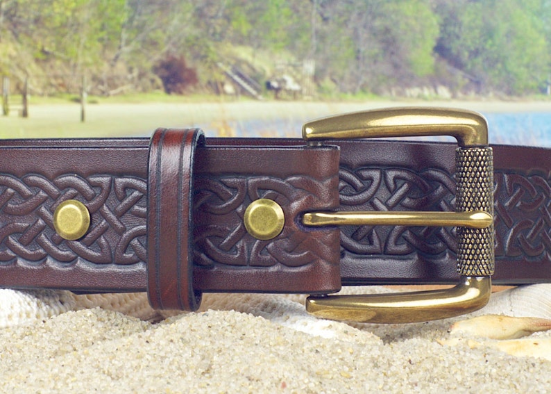 May include: A brown leather belt with a brass buckle and Celtic knot design. The belt has a textured surface and is decorated with brass rivets.