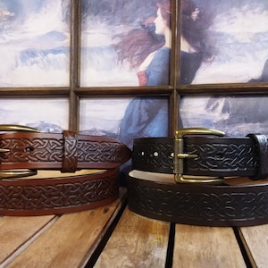 Könnte beinhalten: Zwei Ledergürtel mit keltischen Knotenmustern. Der Gürtel links ist braun und der Gürtel rechts ist schwarz. Beide Gürtel haben messingfarbene Schnallen.