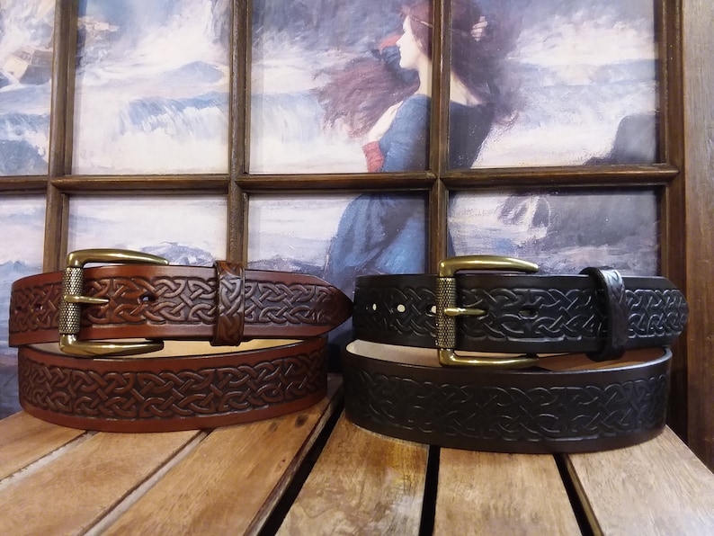 May include: Two leather belts with Celtic knot designs. The belt on the left is brown and the belt on the right is black. Both belts have brass buckles.