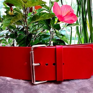 May include: A wide red belt featuring a large, polished silver-tone metal buckle and a matching red leather keeper loop. The belt has a smooth, solid red finish with visible adjustment holes and a rounded tip, presented in a horizontal orientation.