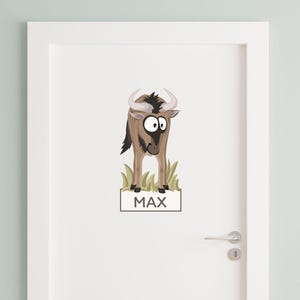 Könnte beinhalten: Ein Wandaufkleber auf einer weißen Tür, der ein Gnu im Cartoon-Stil zeigt, das in grünem Gras steht, über einem rechteckigen Namensschild mit dem Text MAX in schwarzen Großbuchstaben. Das Gnu hat große weiße Augen und geschwungene Hörner.