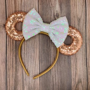 May include: A gold headband featuring sequined ears and a sequined bow. The ears are donut-shaped and covered in copper-coloured sequins. The bow is white with iridescent sequins. The headband is on a wooden surface.