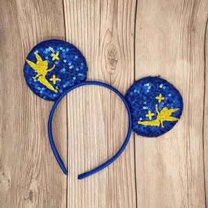 May include: Blue sequin headband with a gold Tinkerbell silhouette and stars. The headband is made of a blue fabric with gold sequins and features a gold Tinkerbell silhouette with a star on each side.