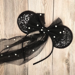 May include: Black headband with a large black bow and two black ears covered in sequins and white pearl accents. The headband is decorated with a black tulle veil.