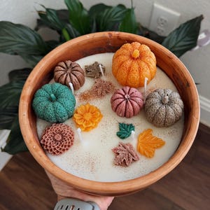 May include: A round terracotta bowl filled with white wax and multiple candle wicks. The surface is decorated with small wax figures including pumpkins in orange, green, pink, and brown, along with orange and yellow flowers and various colored leaf shapes.