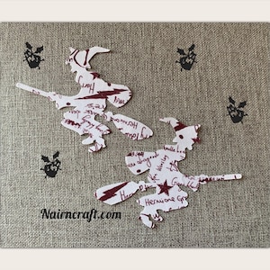 Peut inclure: Deux découpages en papier blanc en forme de sorcières sur des balais, ornés de texte et d'étoiles rouge foncé. Ils sont disposés sur un fond en tissu beige texturé, avec quatre petites silhouettes de chauves-souris noires et le texte Nairncraft.com.