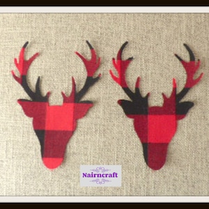 May include: Two flat deer head silhouettes crafted from red and black buffalo plaid fabric. Each deer head features detailed antlers and a distinct checkered pattern. A small rectangular label with the purple text Nairncraft is centred below.